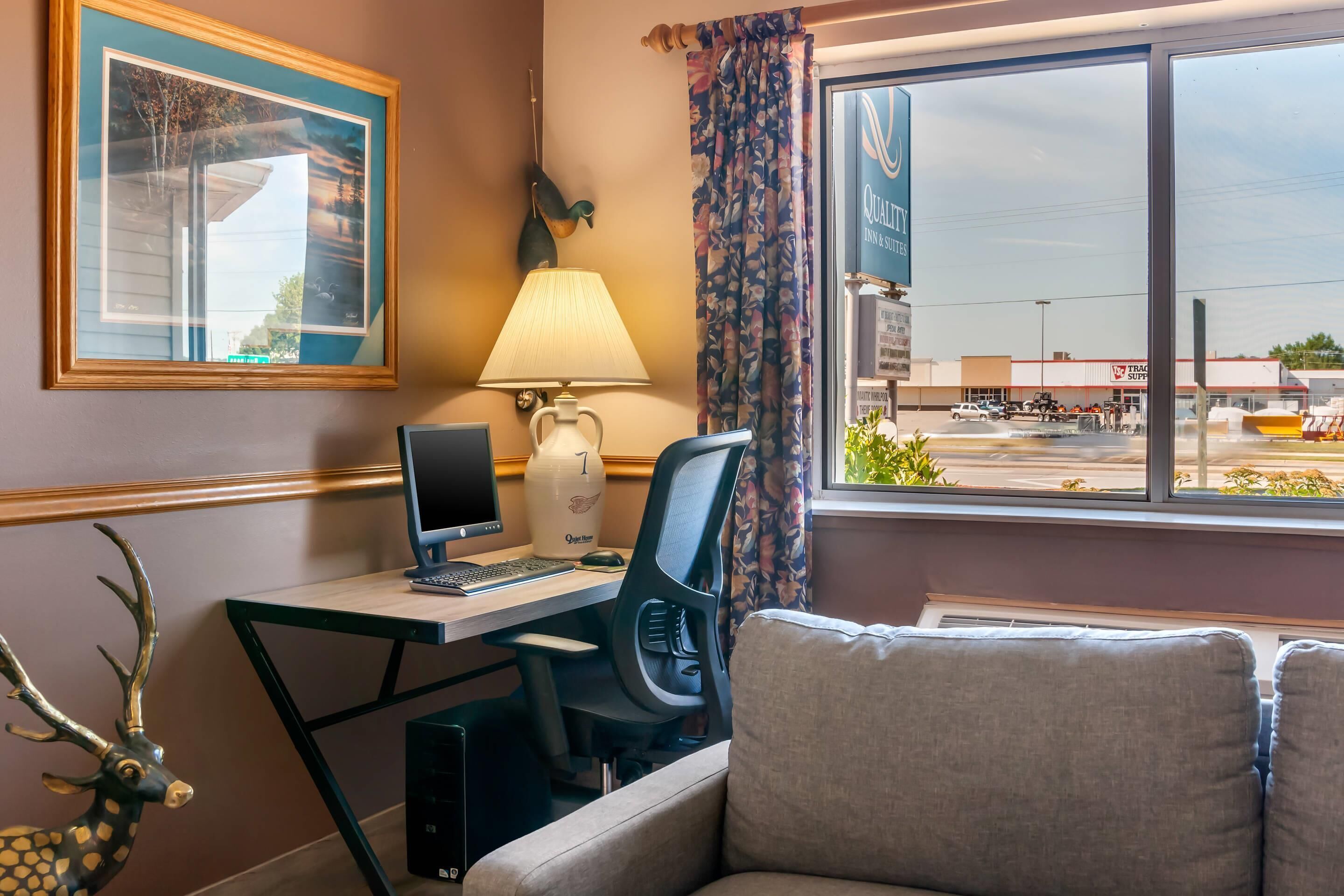 A hotel room with a desk, computer, lamp, and a view of a parking lot.