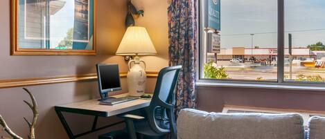 A hotel room with a desk, computer, lamp, and a view of a parking lot.