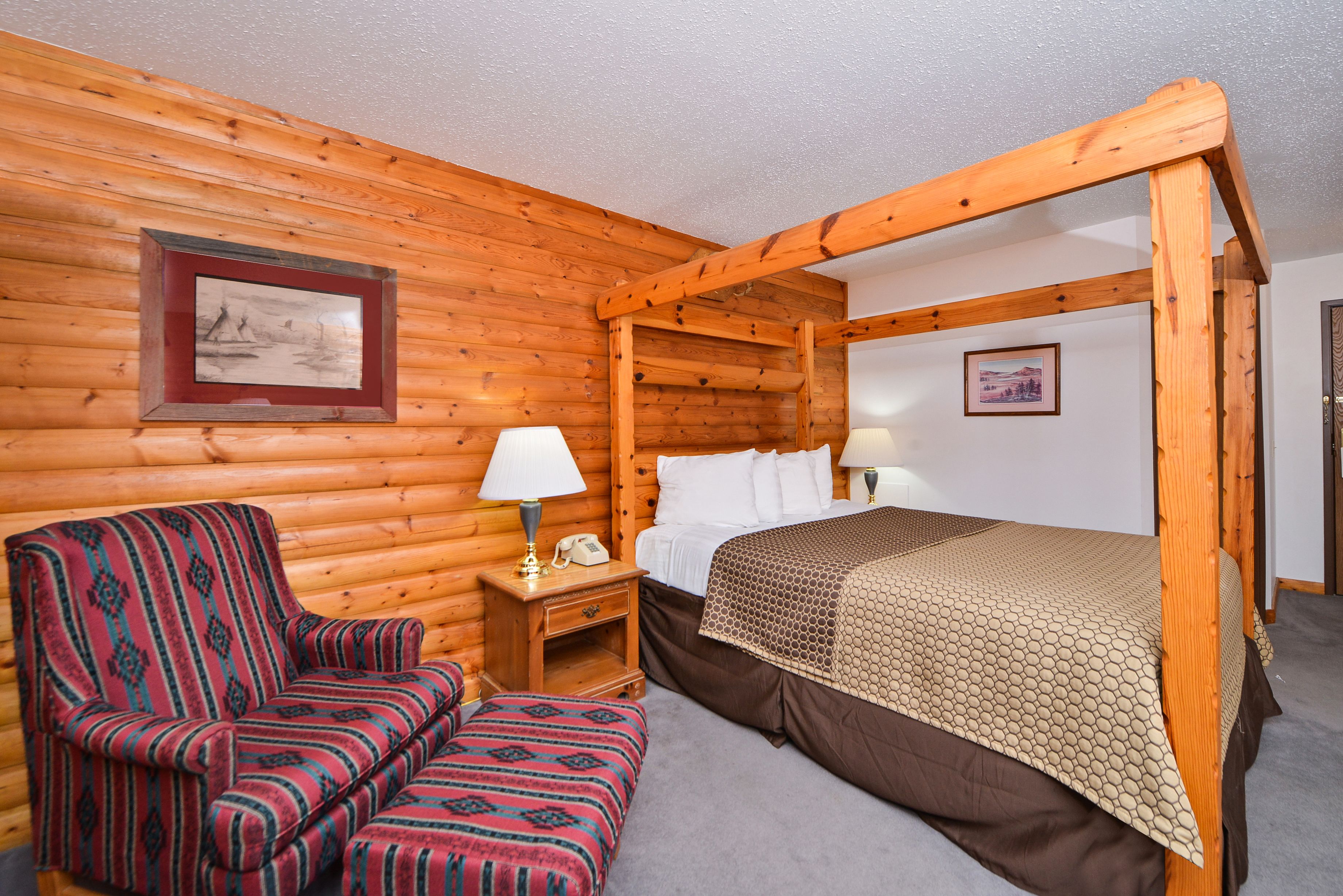 A wooden cabin room with a four-poster bed, a single chair, a nightstand with a lamp, and two framed pictures on the walls.