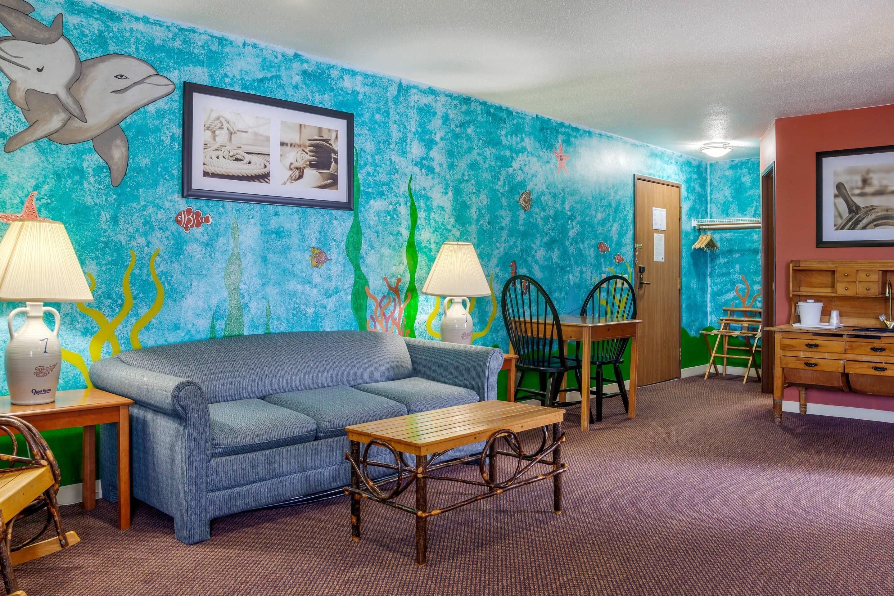 A room with a blue ocean-themed wall, a sofa, a wooden table, a lamp, a chair, and a wooden cabinet.
