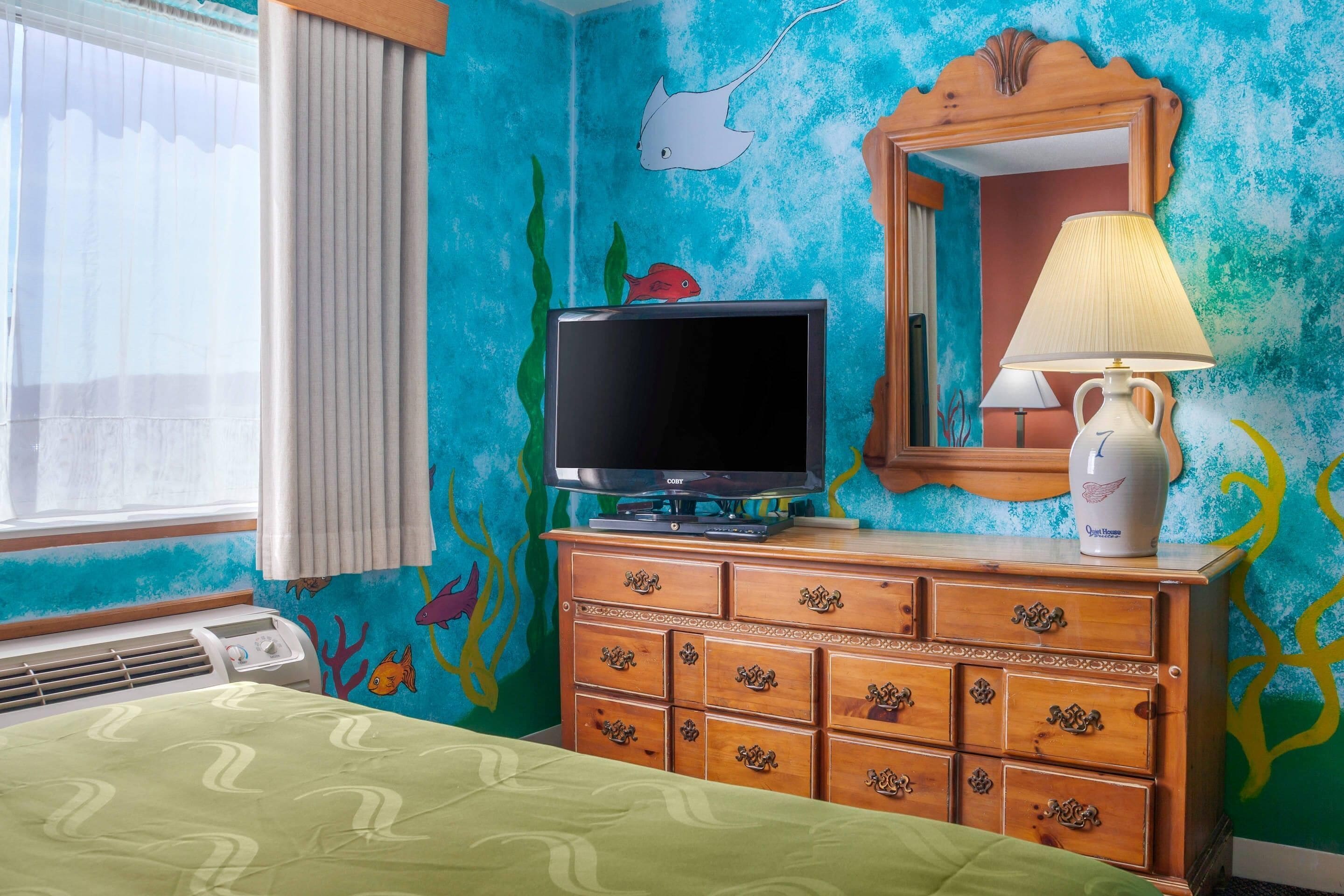 Une chambre avec un lit, un téléviseur posé sur une commode en bois, une lampe et un miroir.