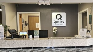 A hotel reception desk with a sign reading "Quality by Choice Hotels," a digital check-in kiosk, a telephone, and a vase with flowers. - Quality Inn & Suites Prairie Du Chien (Prairie Du Chien)