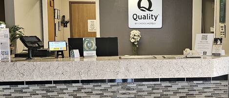 A hotel reception desk with a sign reading "Quality by Choice Hotels," a digital check-in kiosk, a telephone, and a vase with flowers.