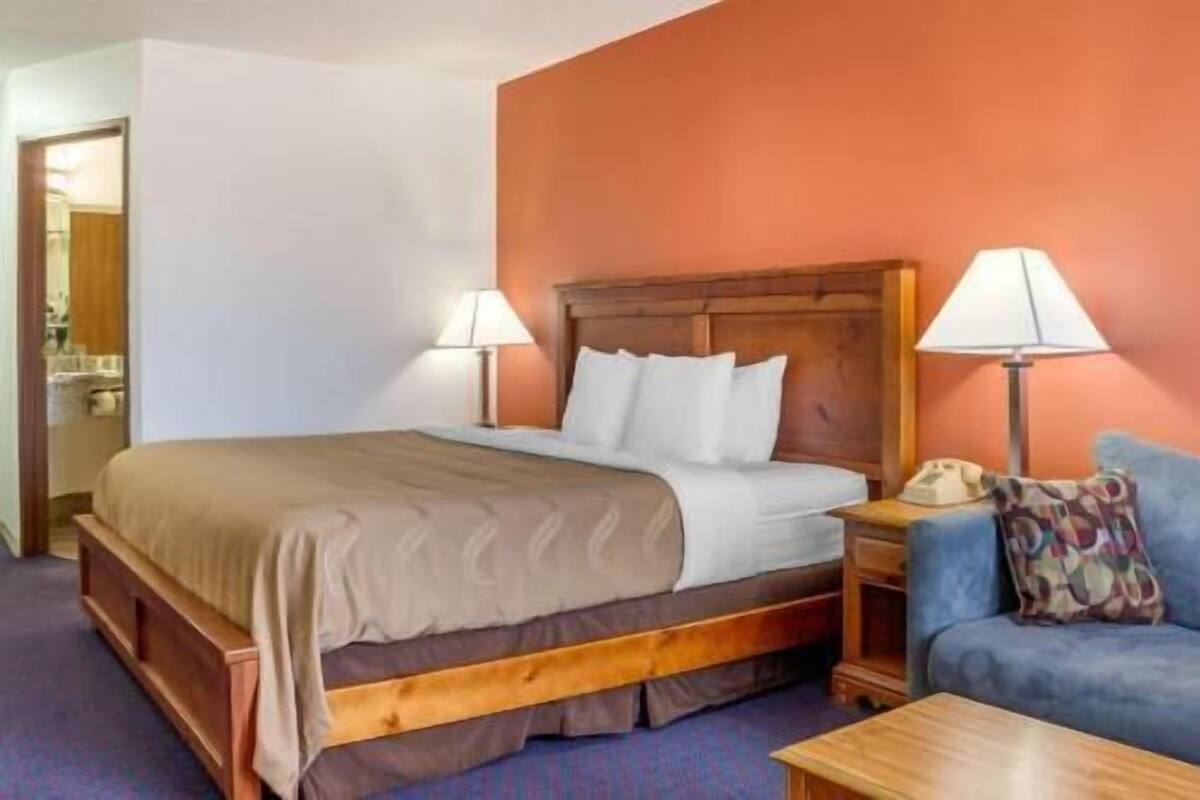 A hotel room with a bed, bedside lamps, a bedside table, a chair, and a wooden headboard.