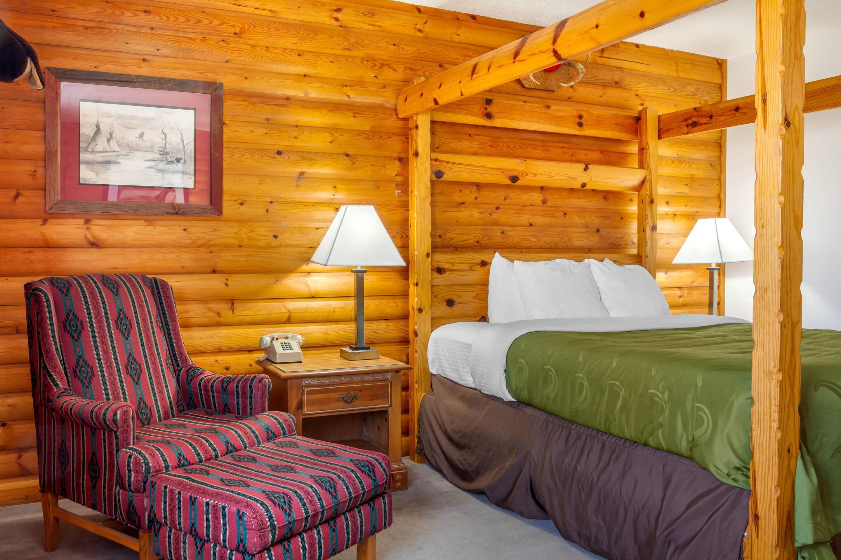 A log cabin room with a bed, a striped armchair, two bedside tables with lamps, and a framed picture on the wall.