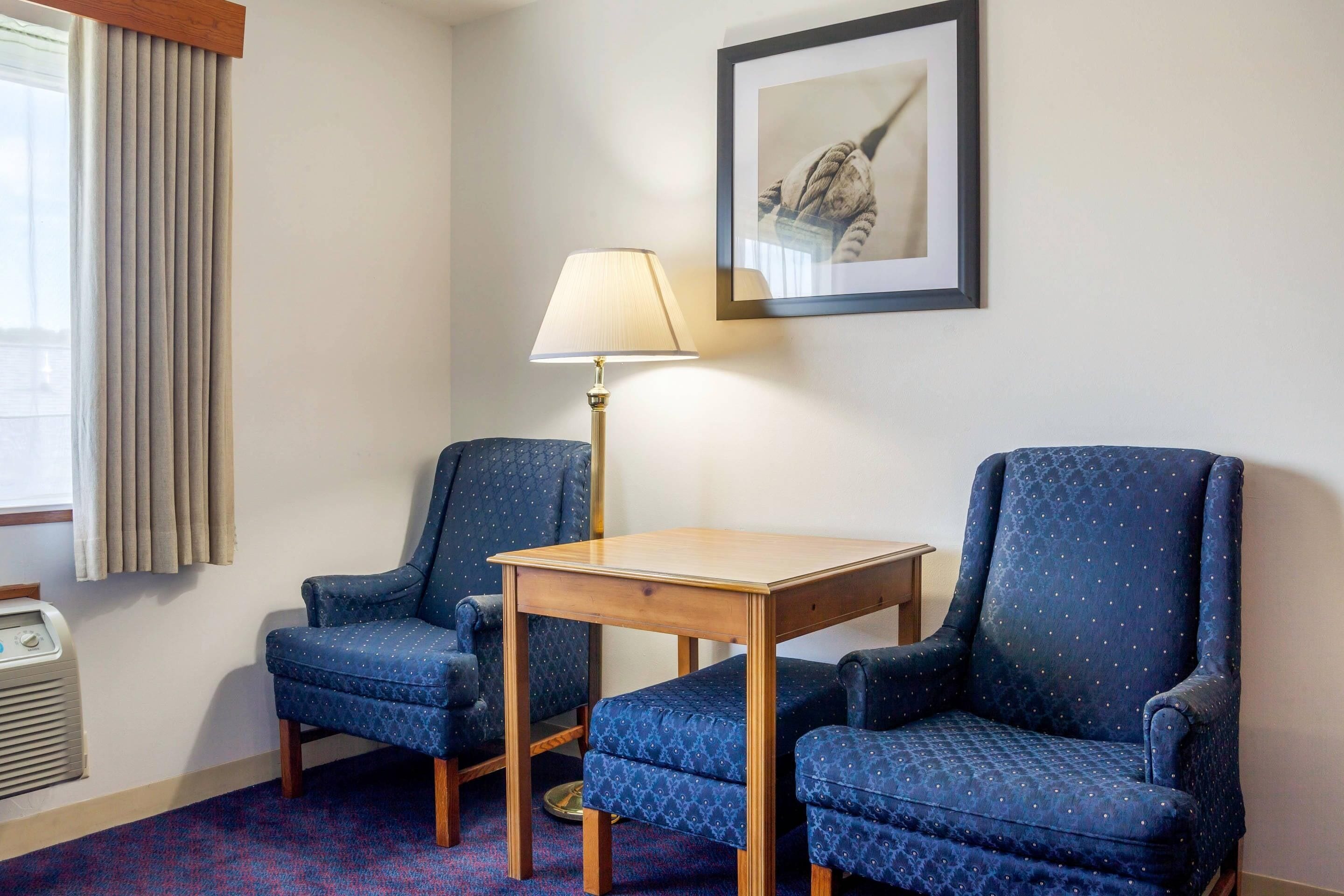 A room with two blue armchairs, a wooden table, a floor lamp, a radiator, and a framed picture on the wall.