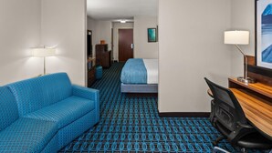 A hotel room with a blue patterned sofa, a bed with a blue blanket, a desk with a lamp, and a framed picture on the wall. - Wingate by Wyndham Oshkosh (Oshkosh)