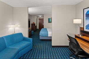 A hotel room with a blue patterned sofa, a bed with a blue blanket, a desk with a lamp, and a framed picture on the wall.
