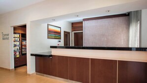 A hotel reception area with a wooden reception desk, a 'Corner Market' sign, and a painting on the wall.