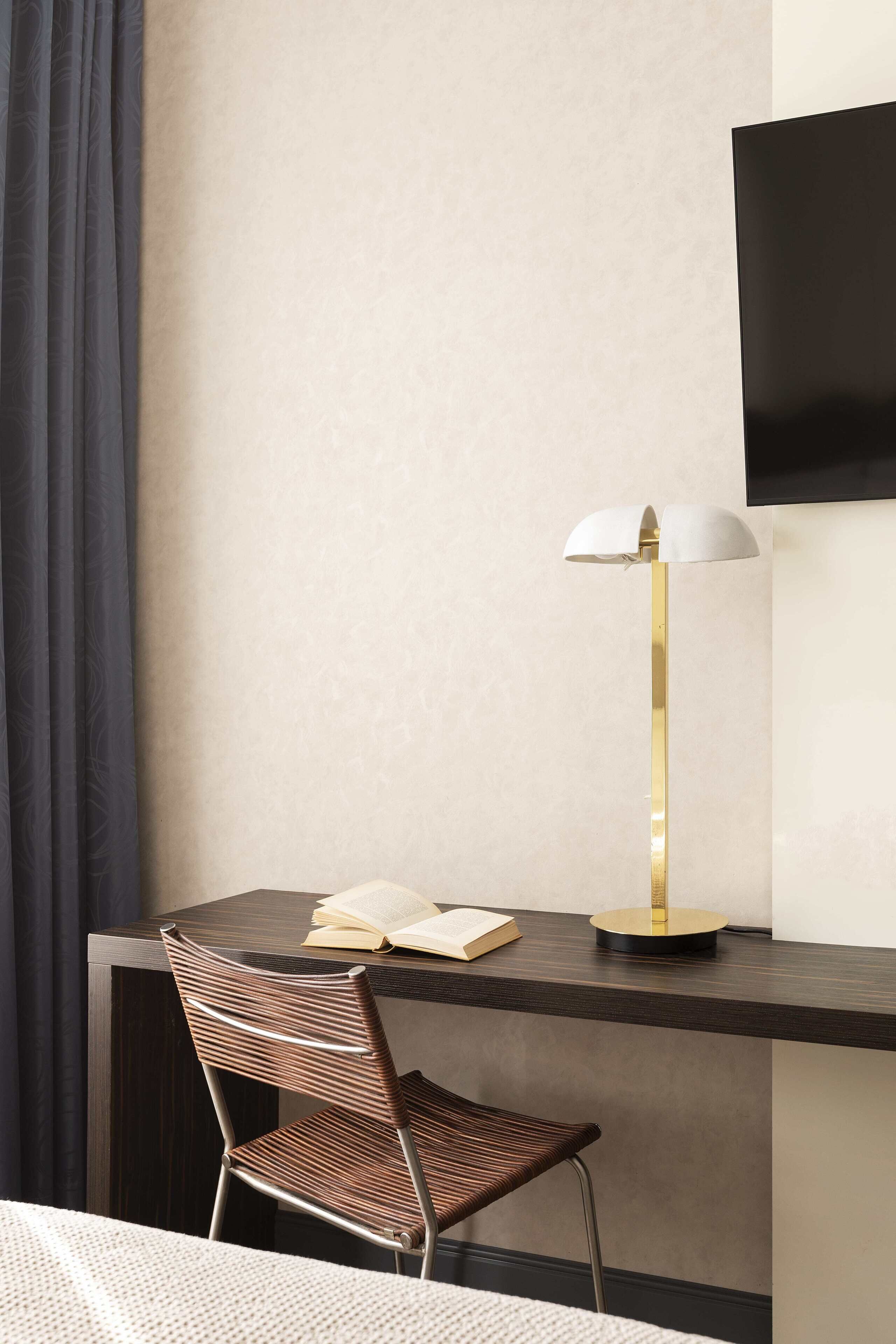 Habitación de hotel moderna con un escritorio de madera oscura, una sola silla, una lámpara de pie y un televisor de pantalla plana montado en la pared.