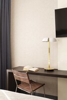 Quarto de hotel moderno com escrivaninha de madeira escura, uma cadeira, abajur e televisão de tela plana presa à parede.