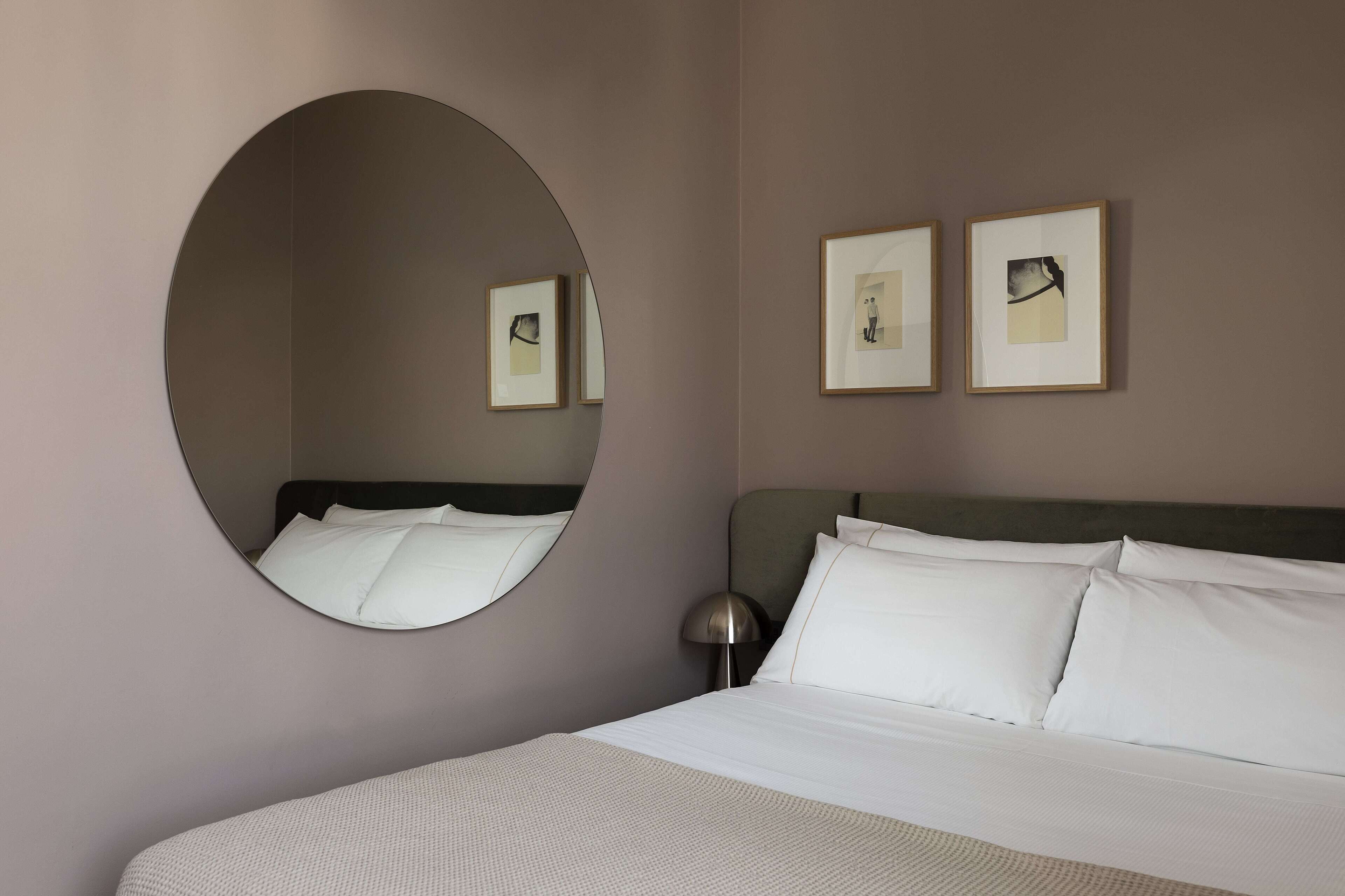Une chambre d’hôtel avec un grand miroir rond, un lit recouvert de draps blancs et deux tableaux encadrés accrochés au mur.