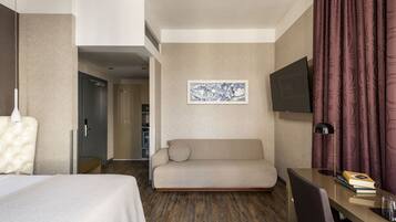 A hotel room with a bed, a sofa, a desk with a chair, a TV, and a painting on the wall.