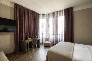 A hotel room with a bed, a desk with a chair, a TV, and large windows with curtains.