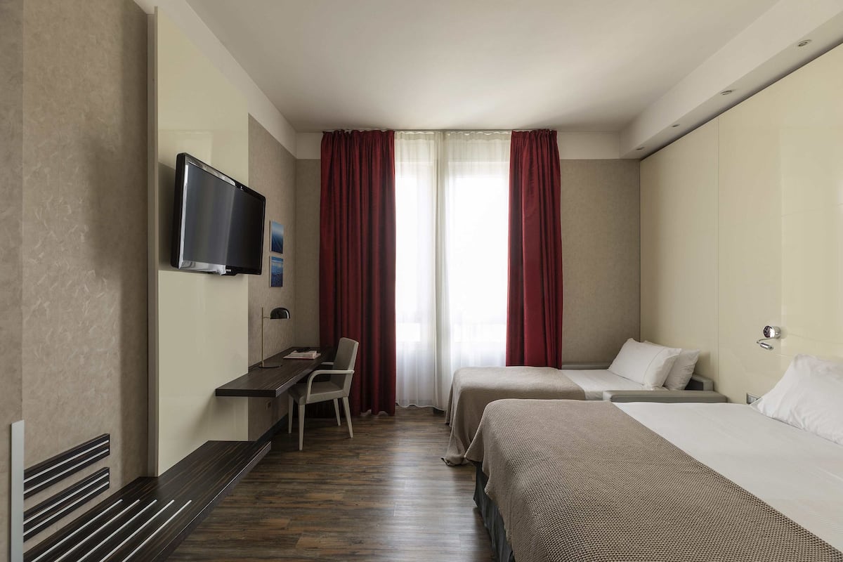 A hotel room with a flat-screen TV mounted on the wall, a desk with a chair, two beds, and a window with red curtains.