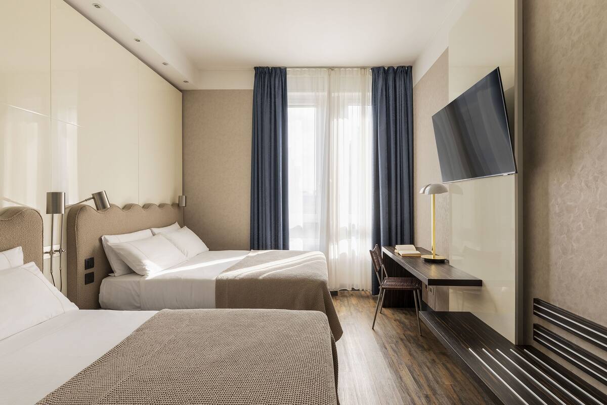 A hotel room with two beds, a desk, a TV, and a window with curtains.