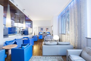Un soggiorno moderno con arredamento bianco e blu, un'ampia finestra e un angolo cottura sullo sfondo.