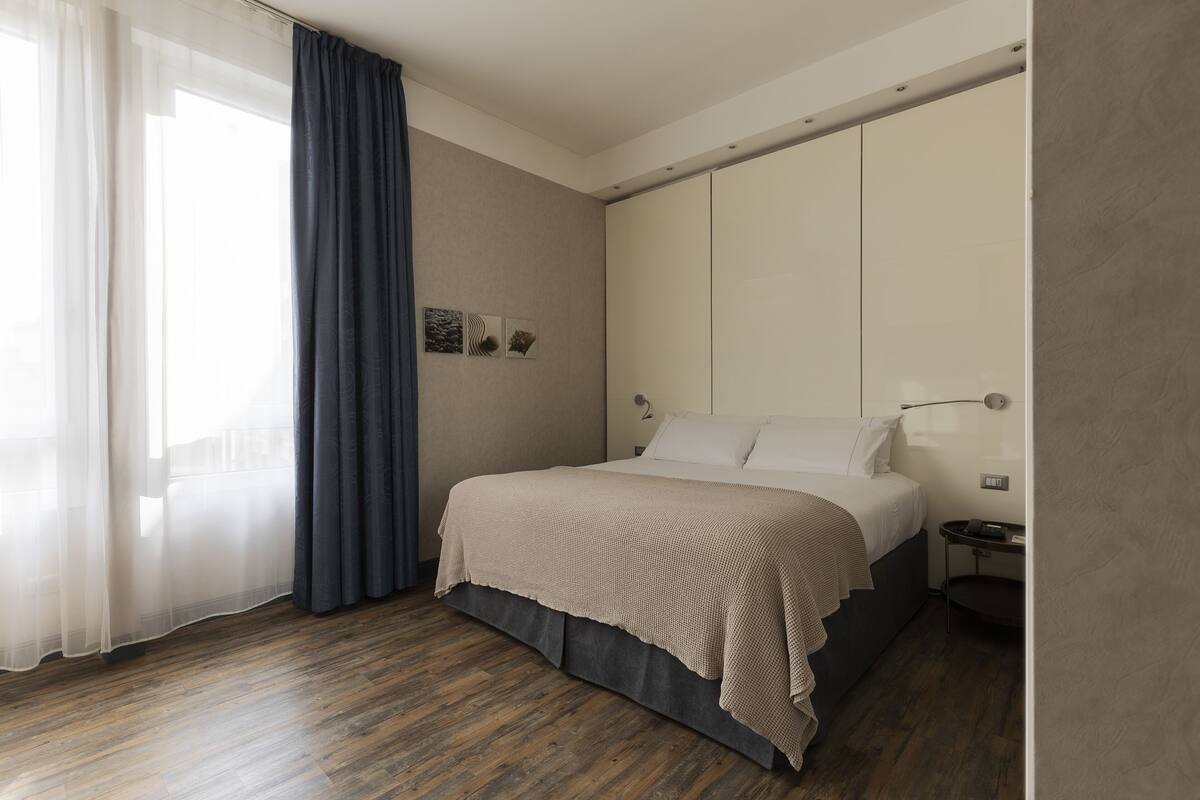 A modern hotel room with a large bed, a bedside table, a nightstand, and a window with sheer curtains.