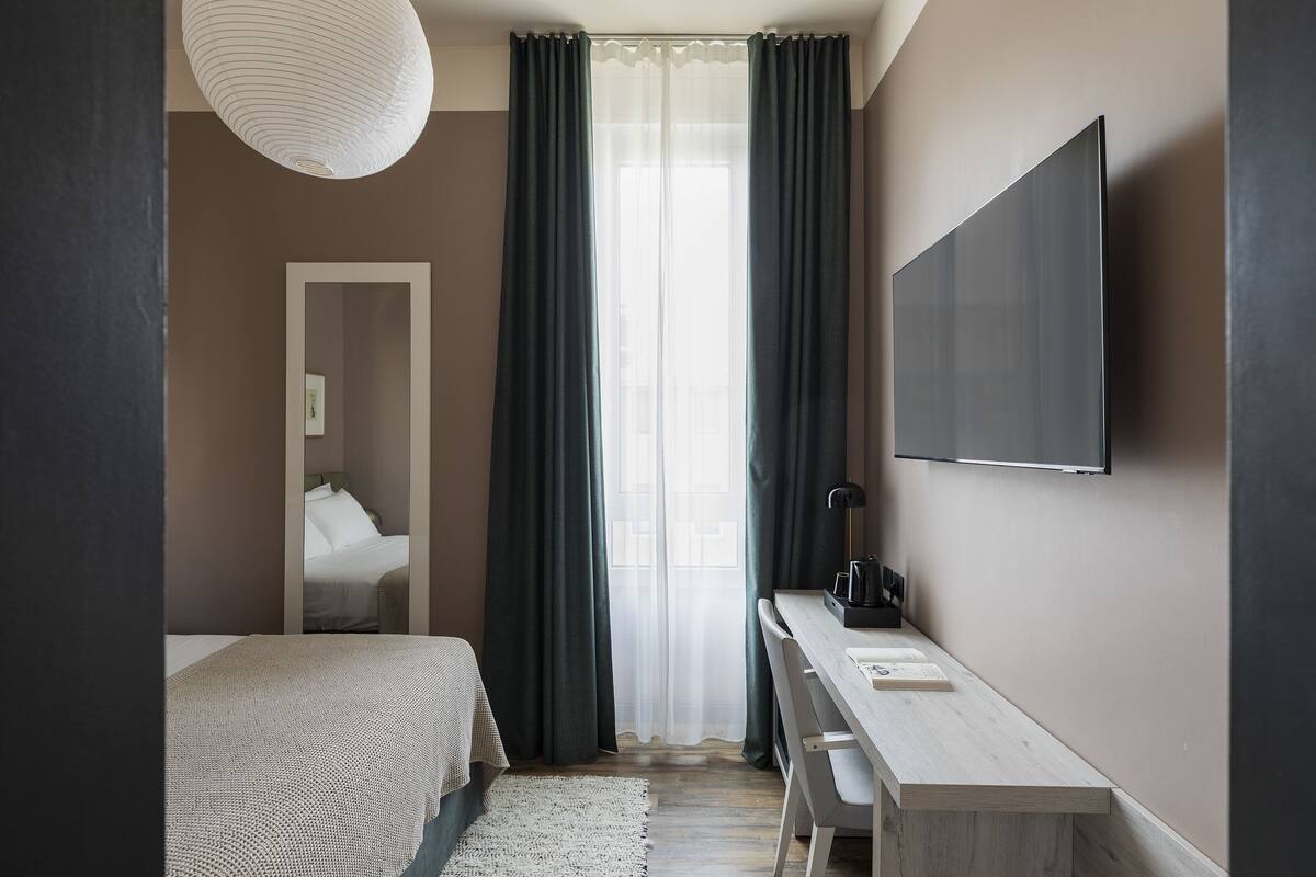 A modern hotel room with a bed, a desk, a TV, and a large window with curtains.