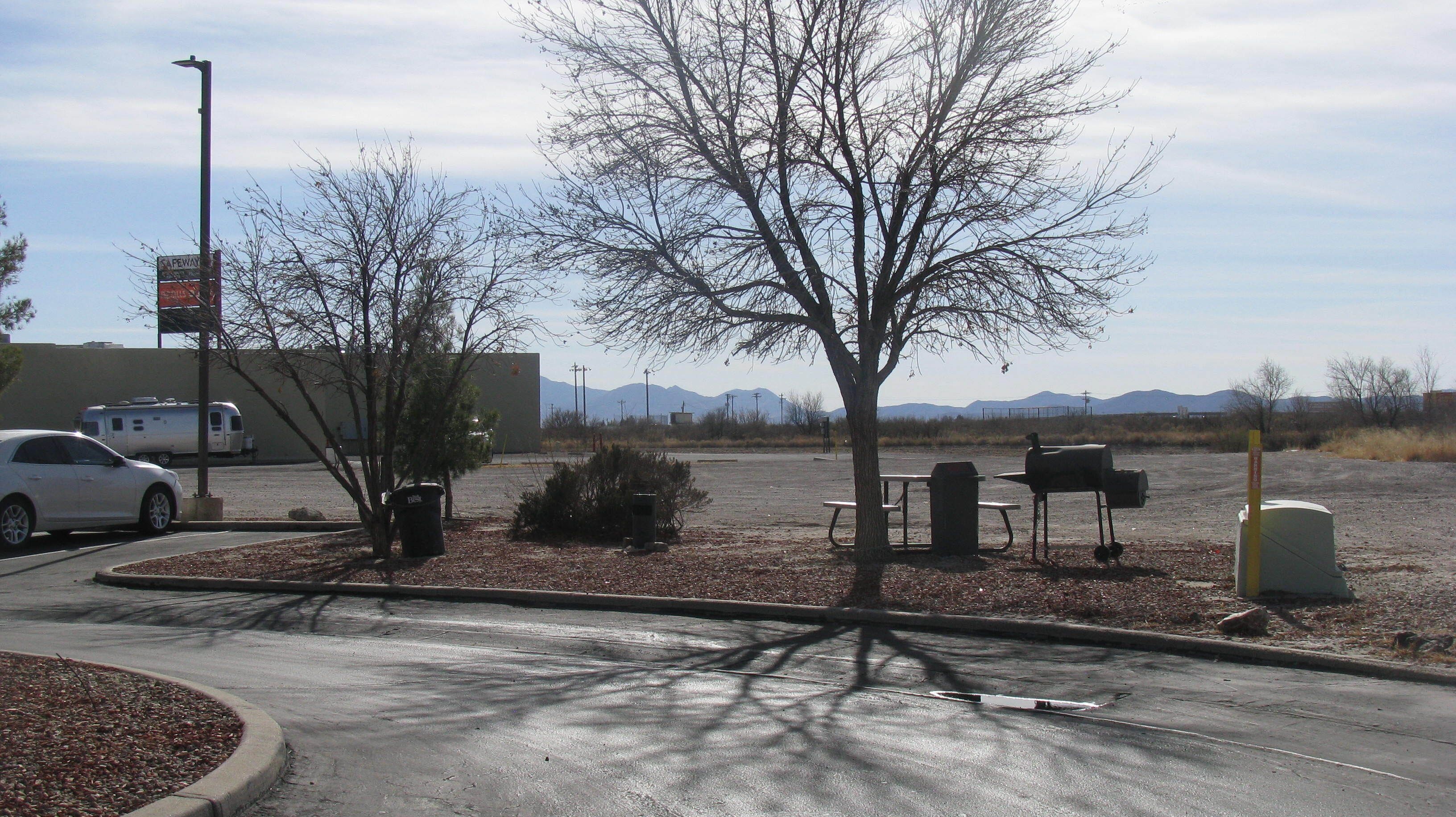 A parking lot with a white car, a bench, a mailbox, and a trash can.