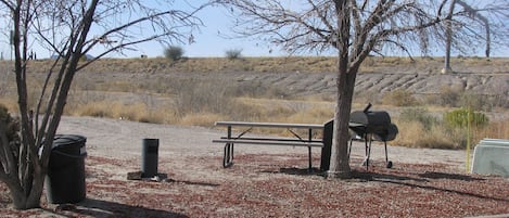 A picnic area with a bench, a barbecue grill, and a trash can.
