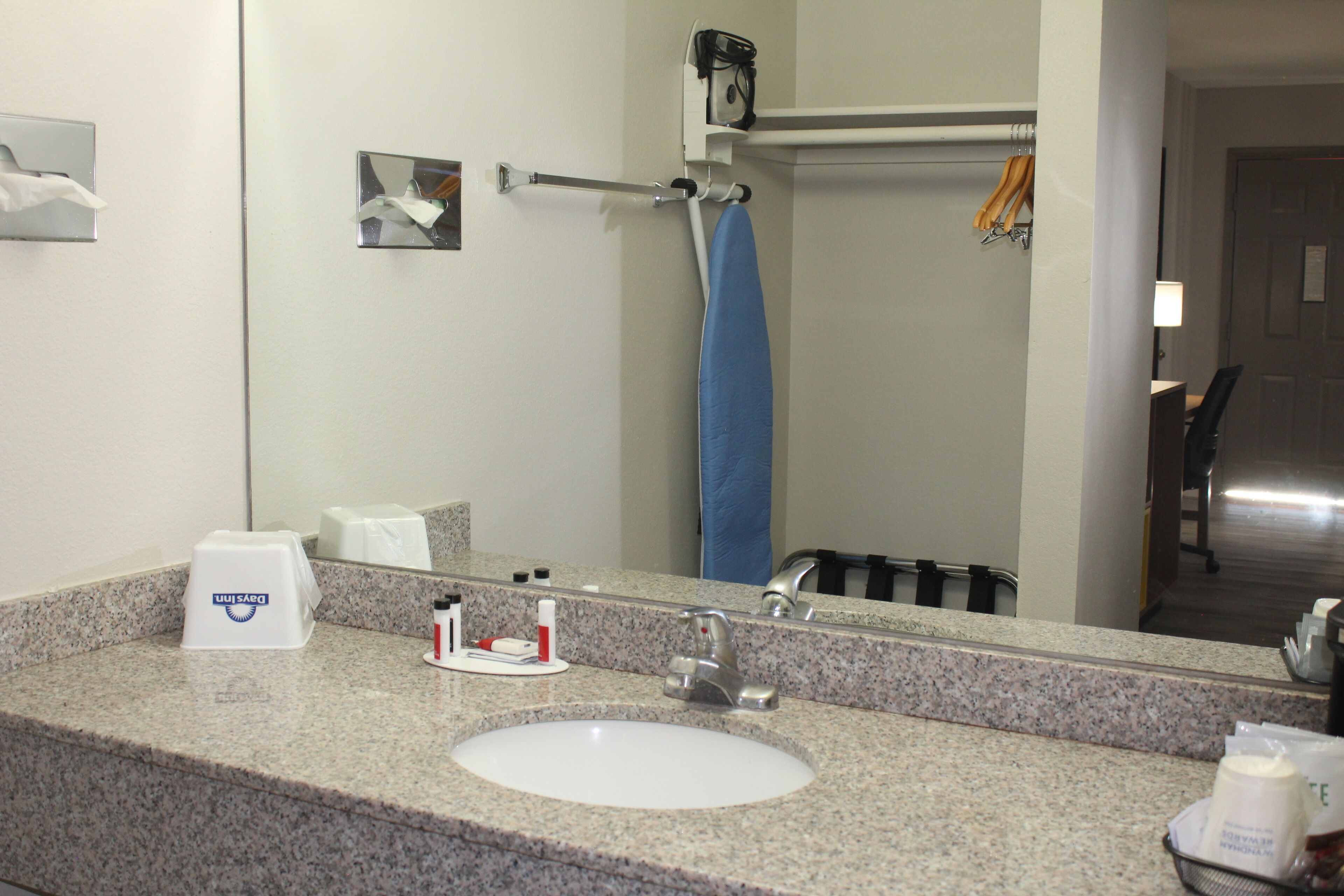 A hotel bathroom with a granite countertop, a white sink, a mirror, a towel rack, and a shower area visible in the reflection.