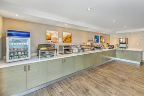 A modern breakfast area with a self-service refrigerator, coffee machines, and a variety of food items.