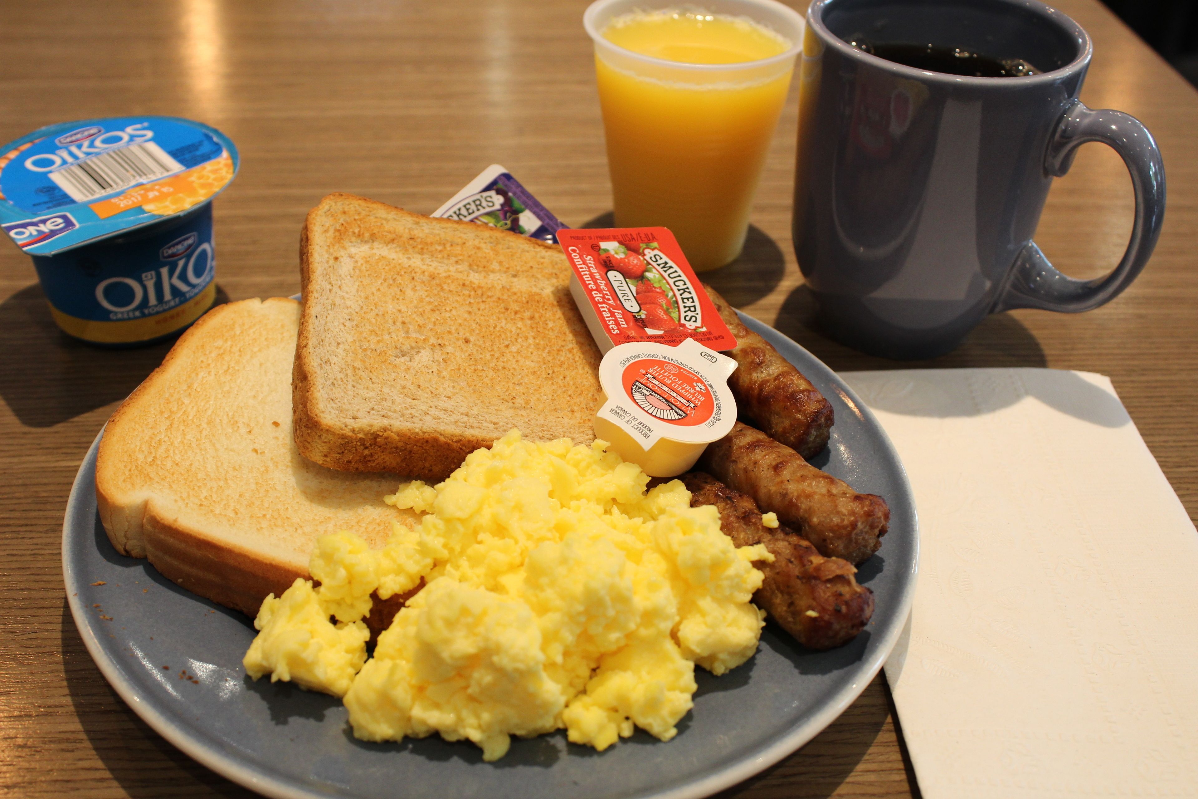 A plate with toast, scrambled eggs, sausages, a cup of coffee, a glass of orange juice, and a container of yogurt.