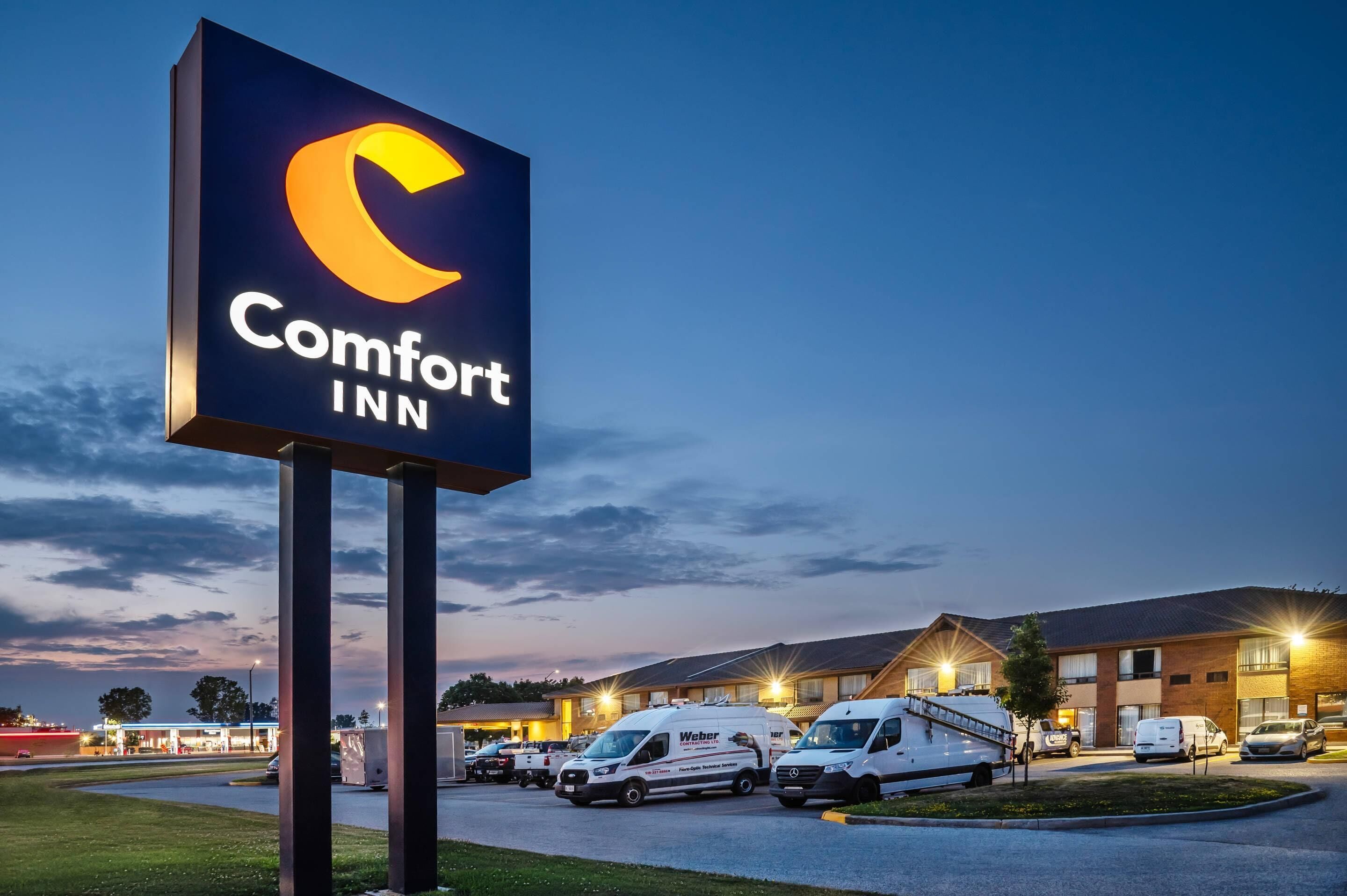 Ein Comfort Inn Schild mit dem Halbmond-Logo, umgeben von einem Parkplatz mit mehreren weißen Lieferwagen und einem Gebäude mit Backsteinfassade.
