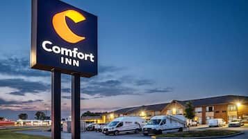 Letrero de Comfort Inn con un logotipo de luna creciente, rodeado por un estacionamiento con varias camionetas blancas y un edificio de fachada de ladrillo.