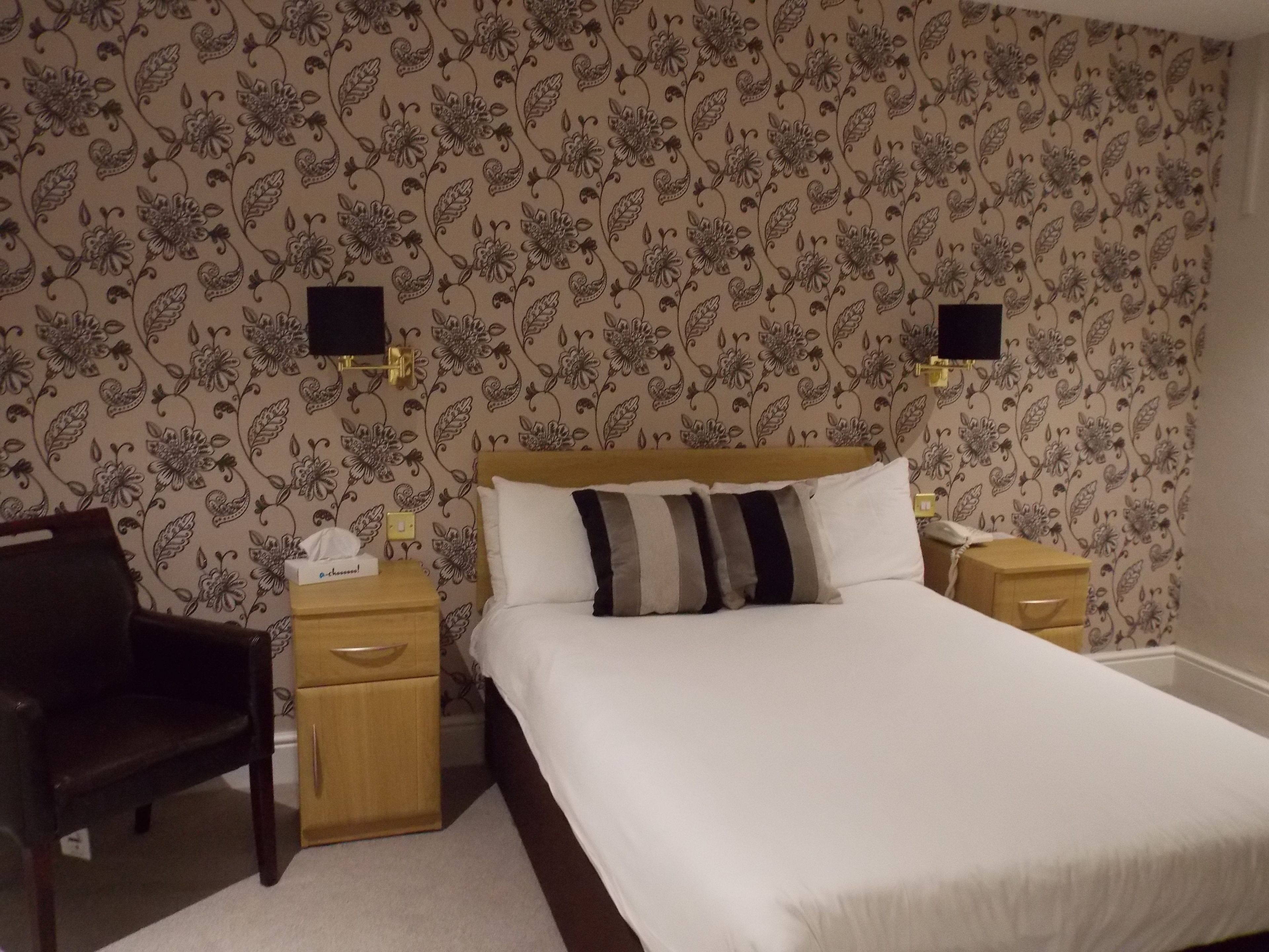 Ein Schlafzimmer mit einem großen Bett, zwei Nachttischen, einem Beistelltisch, einem Holzstuhl und einer blumigen Wandgestaltung.