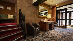 A hotel lobby with a staircase, a leather armchair, a desk with drawers, a lamp, and a large mirror.