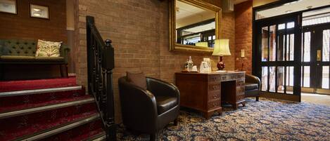 A hotel lobby with a staircase, a leather armchair, a desk with drawers, a lamp, and a large mirror.