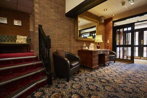 Un vestíbulo de hotel con una escalera, un sillón de cuero, un escritorio con cajones, una lámpara y un espejo grande.