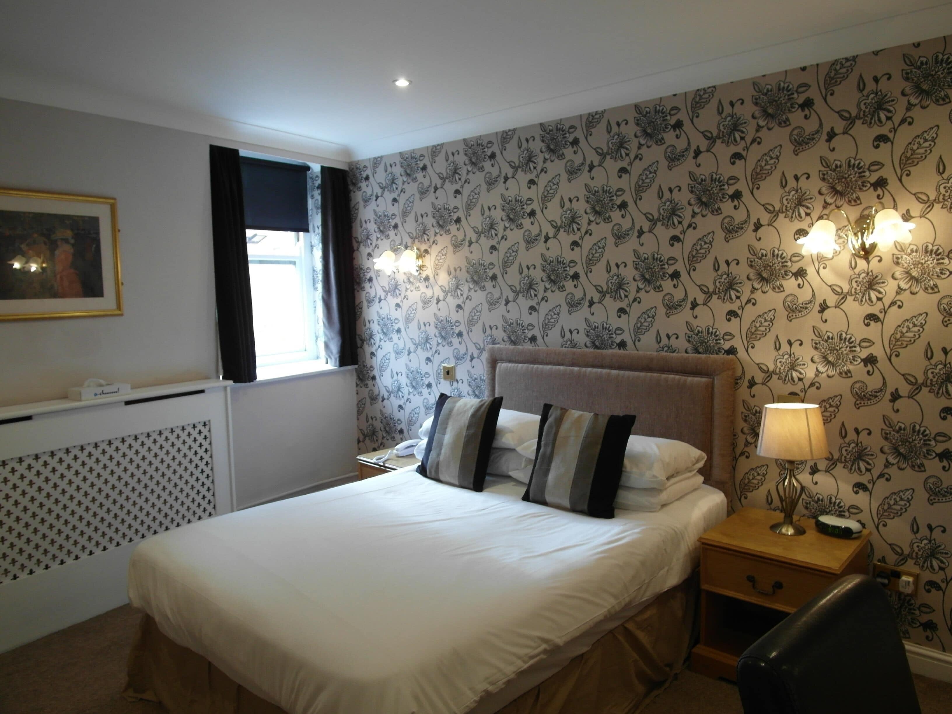 A neatly arranged bedroom with a large bed, a bedside table with a lamp, and a wall with floral wallpaper.
