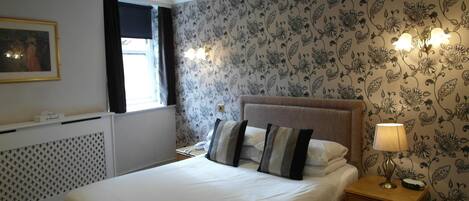 A neatly arranged bedroom with a large bed, a bedside table with a lamp, and a wall with floral wallpaper.