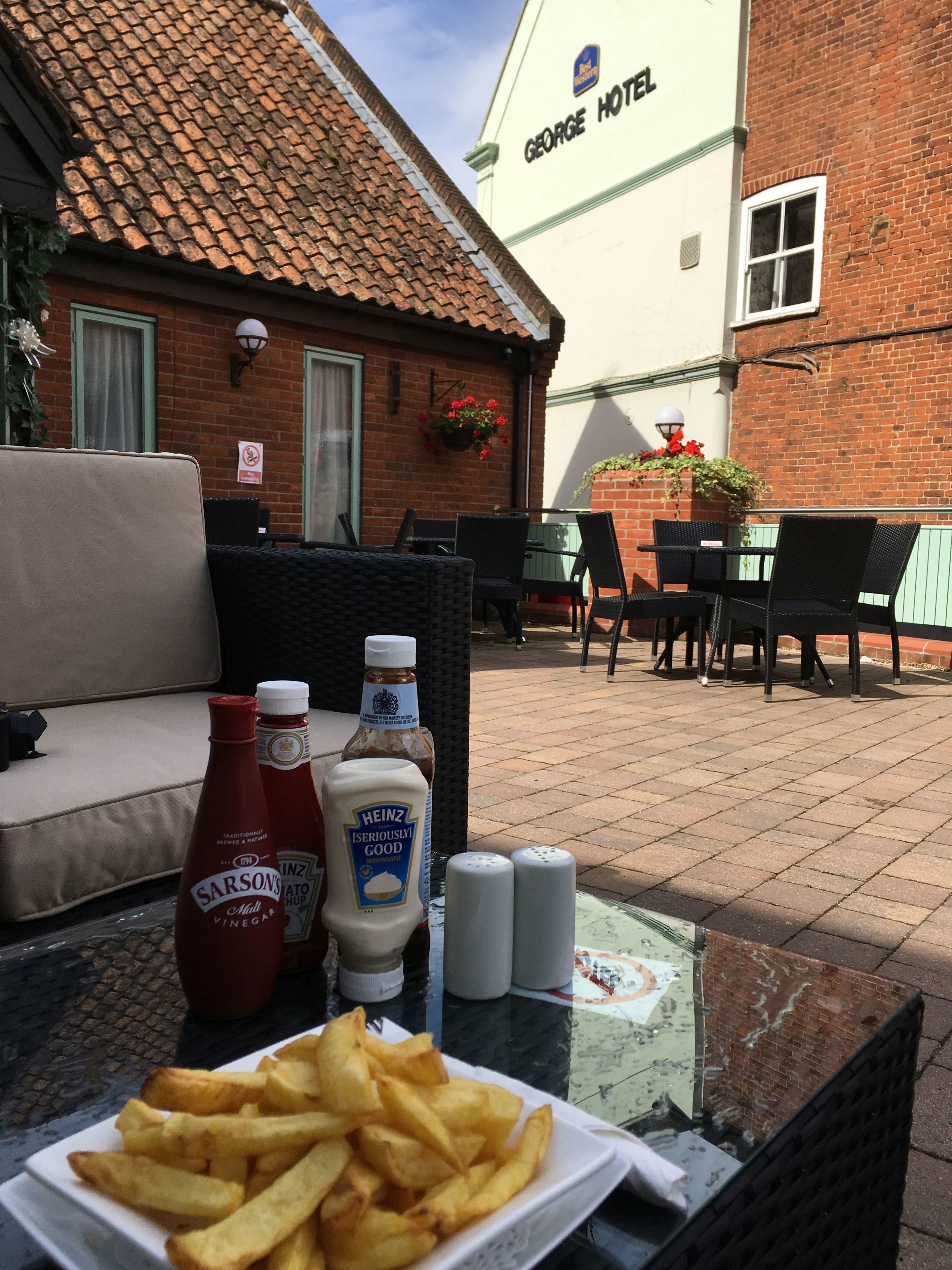 A table with a plate of French fries, bottles of condiments, and salt and pepper shakers outside a building with a sign reading "GEORGE HOTEL".