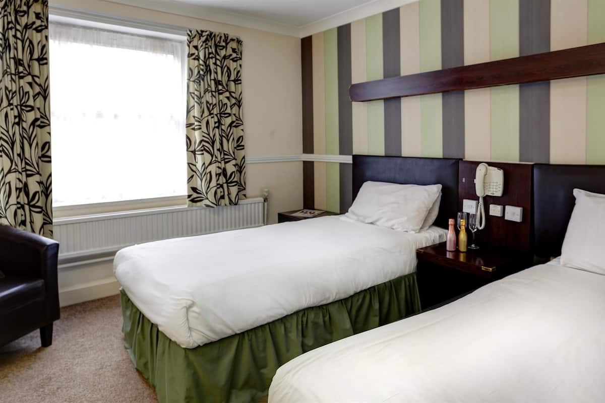 Une chambre d’hôtel avec deux lits simples, une tête de lit en bois, une table de chevet avec une lampe et une fenêtre avec des rideaux.