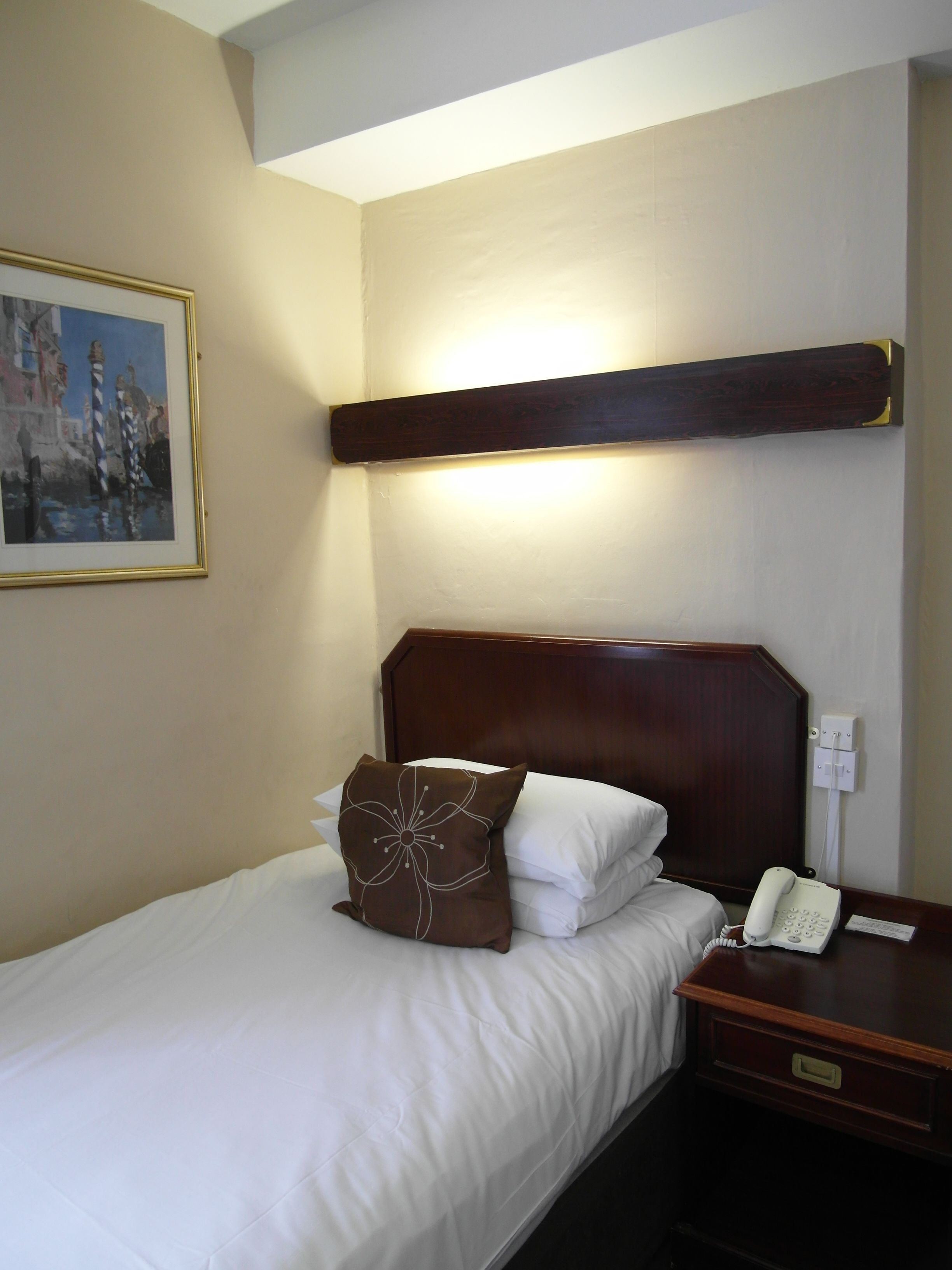 A hotel room with a bed, a nightstand with a telephone, a painting on the wall, and a wooden headboard.