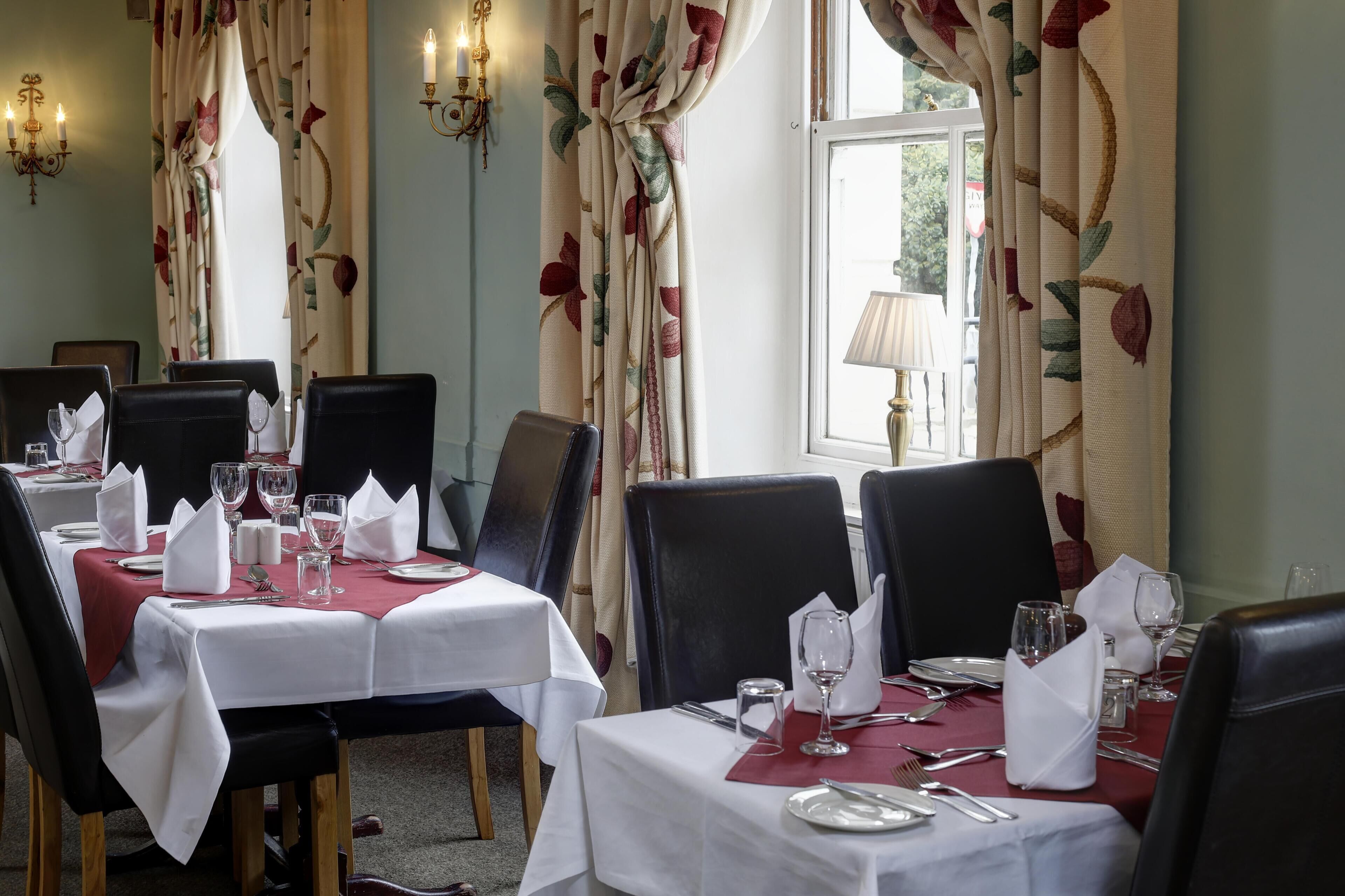 A dining area with tables set for four, each with white tablecloths, black chairs, and neatly arranged cutlery and glassware.