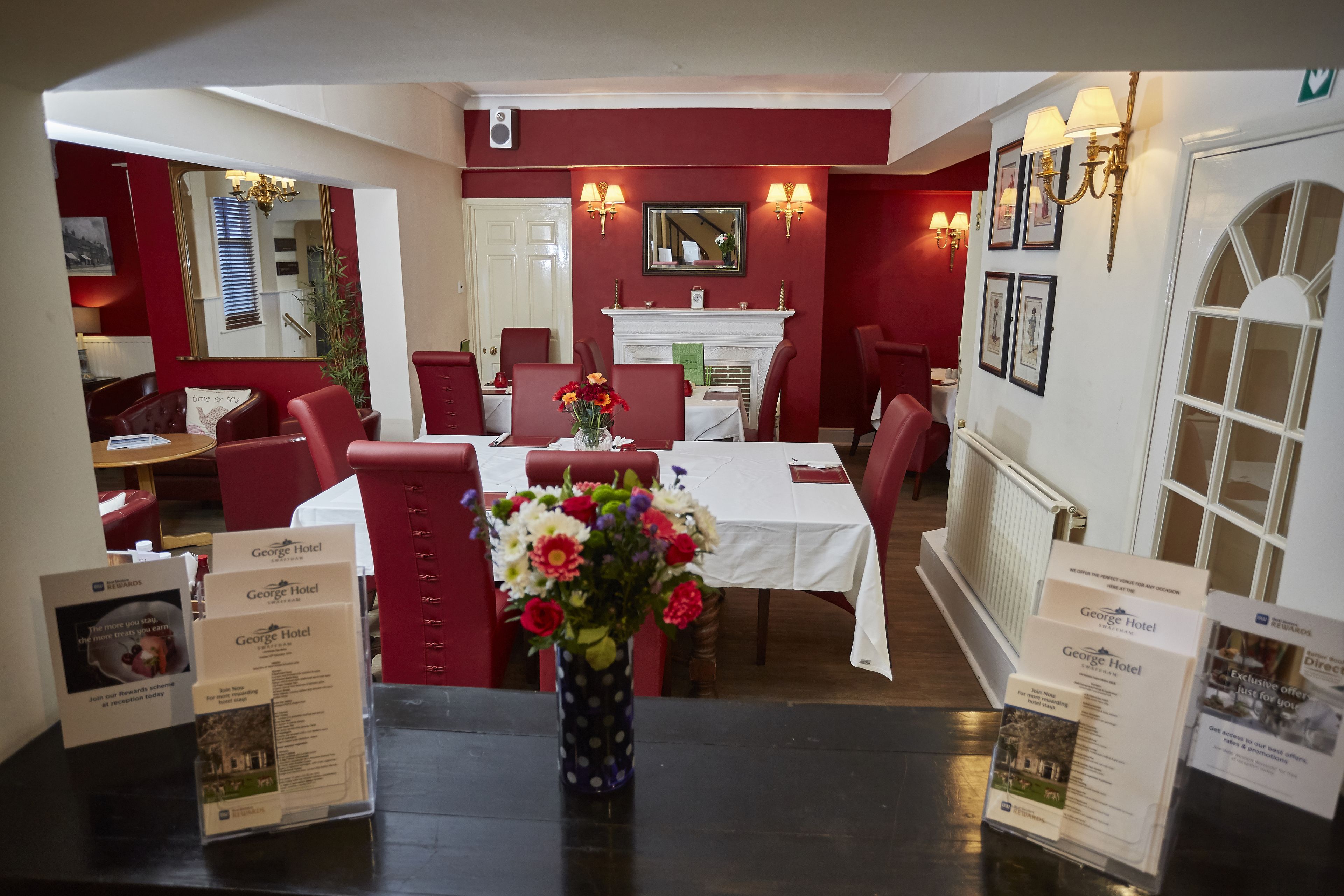 A dining area with red upholstered chairs, a white table, and a vase of flowers. There are brochures on the table and a fireplace in the background.