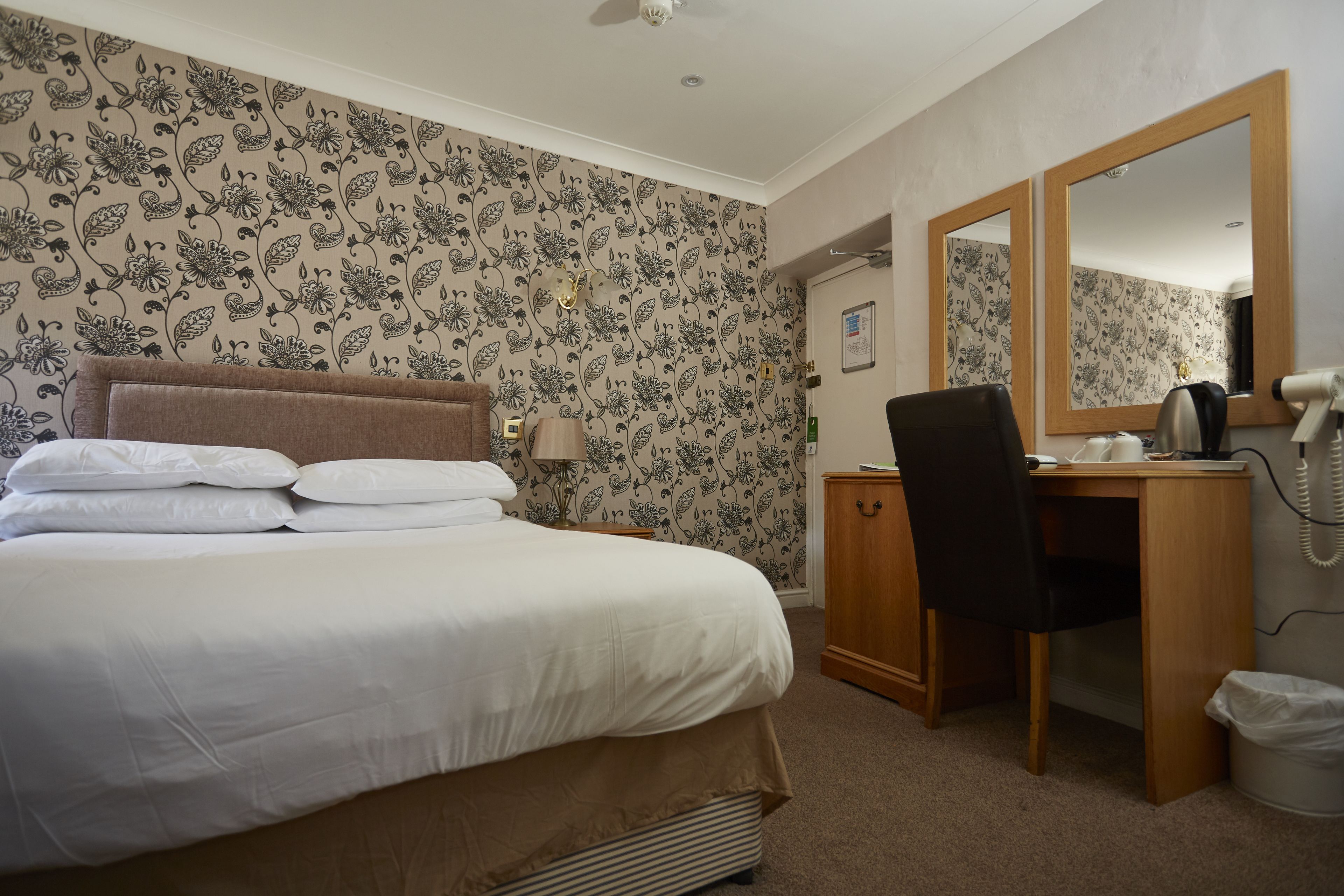 A hotel room with a bed, a desk, a chair, a mirror, and a wall with floral wallpaper.