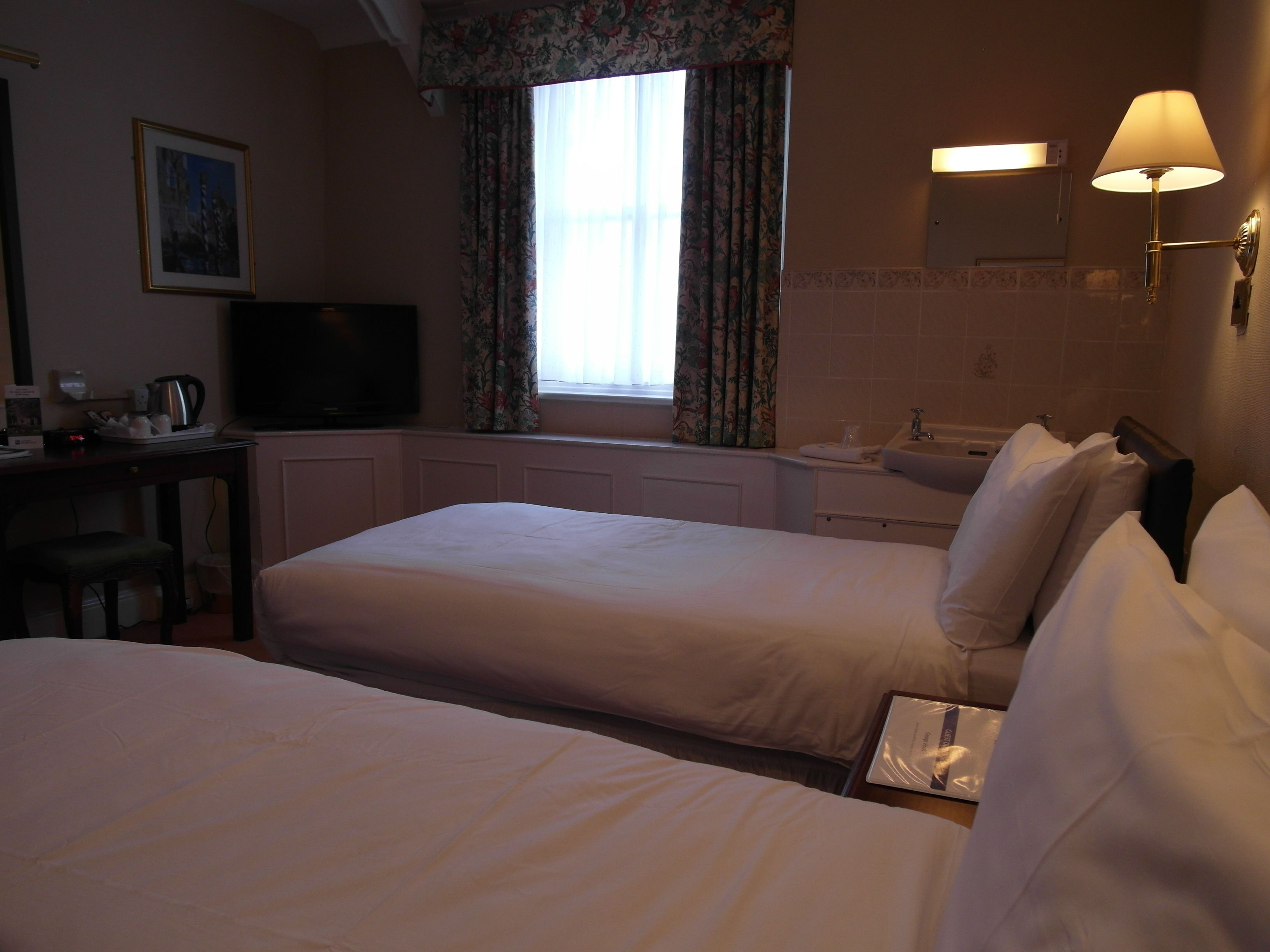 A hotel room with two beds, a desk with a television, a window with curtains, a sink, and a lamp.