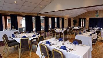 A banquet hall with multiple tables set for dining, featuring white tablecloths, gold chairs, and blue napkins.