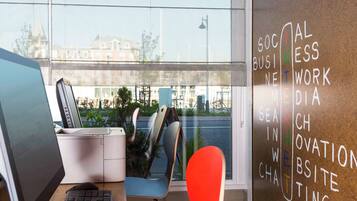 A modern office space with a desk, computer, and a red chair. A window with a view of buildings and greenery is visible.
