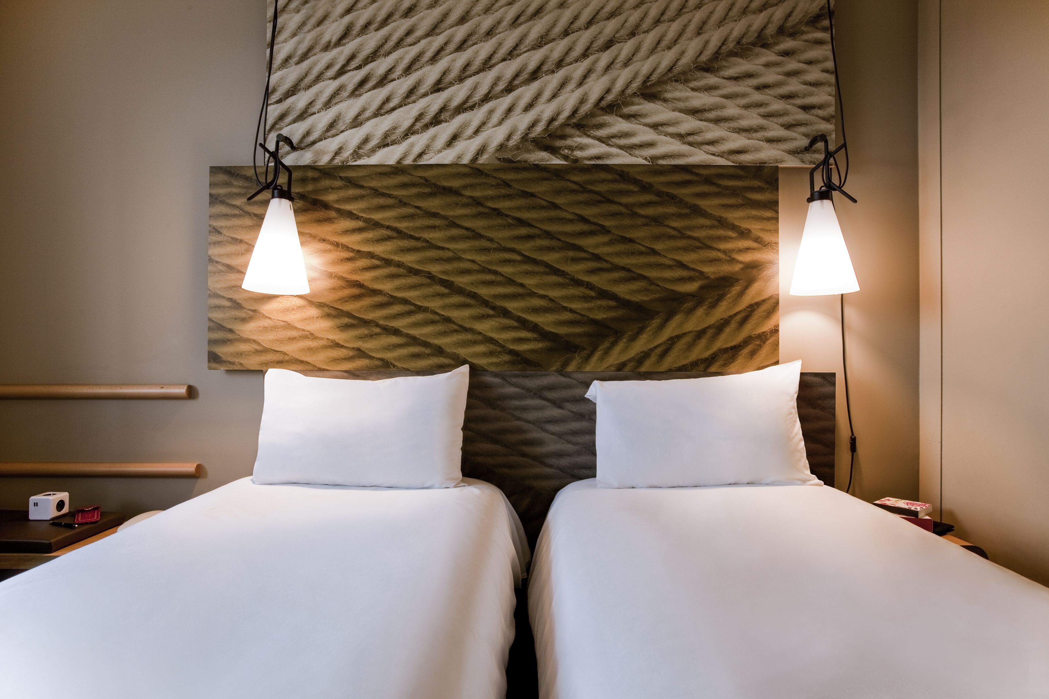 Habitación de hotel con dos camas, cabecero de madera, mesitas de noche y una lámpara fijada a la pared.