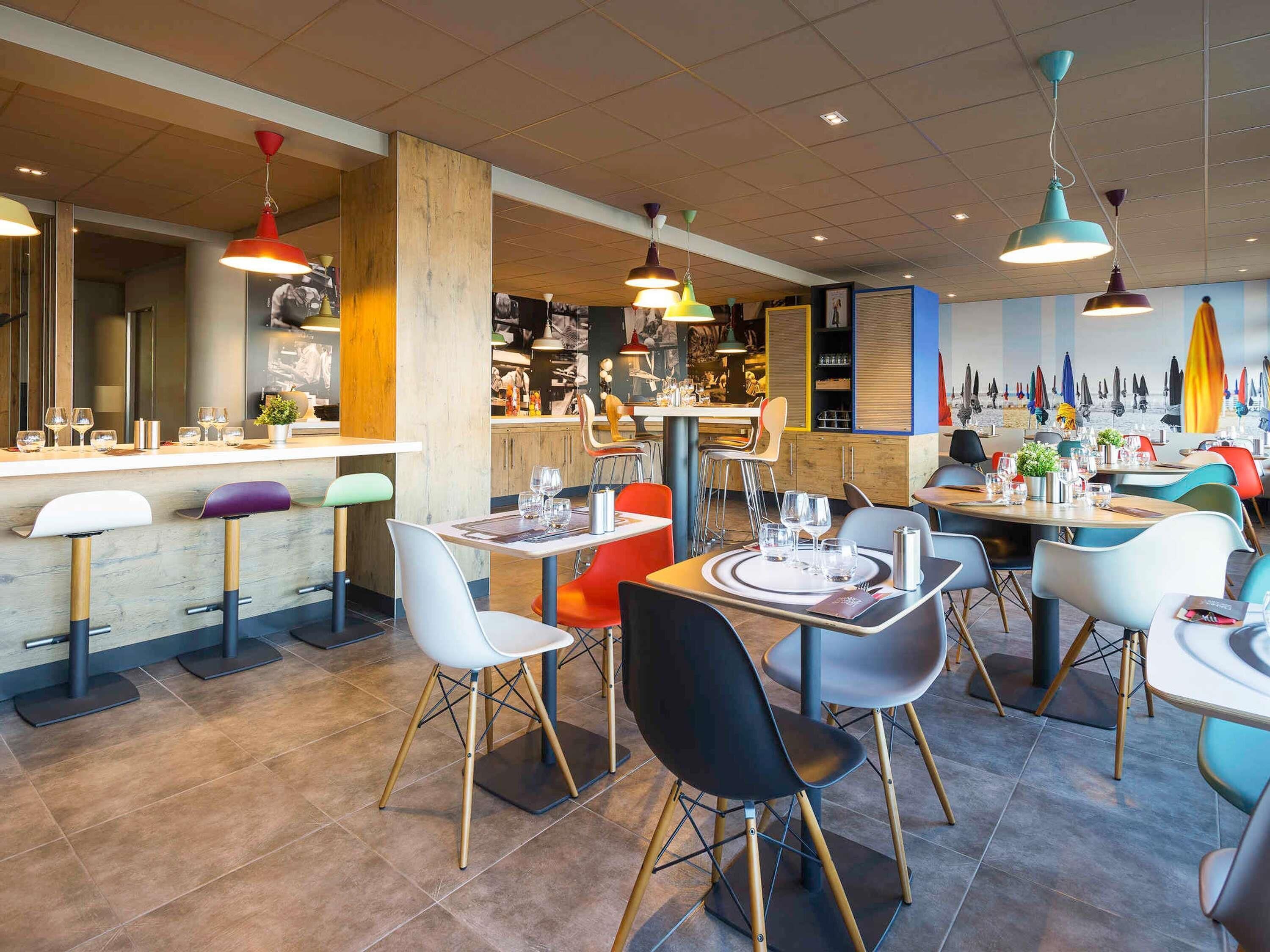 A modern restaurant interior with multiple dining tables, colorful chairs, and pendant lighting.