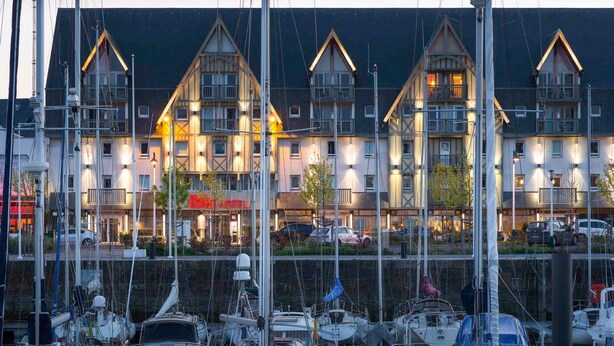 Un port de plaisance où sont amarrés de nombreux bateaux, un hôtel avec une enseigne rouge bien visible, et un bâtiment doté d’un toit triangulaire distinctif.