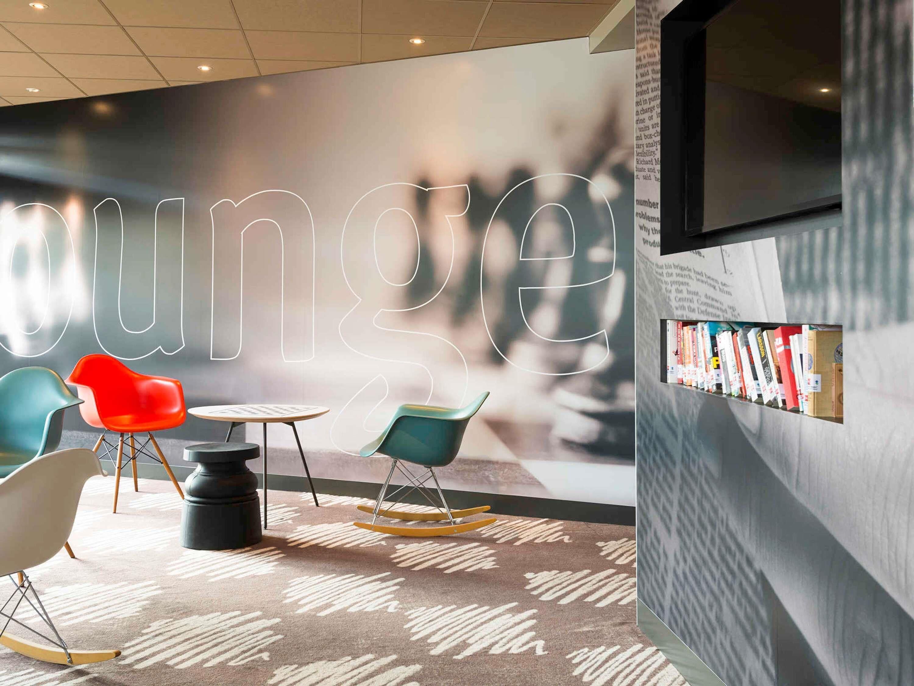 Uma área de estar moderna com um grande letreiro iluminado 'lounge', assentos confortáveis, uma pequena mesa redonda e uma estante com livros.
