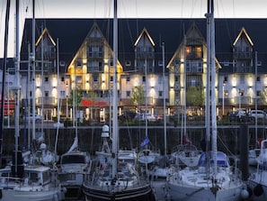 A marina with multiple boats docked, a hotel building with a sign reading 'ibis HOTEL' in the background, and a clear sky. - ibis Deauville Centre (Deauville)